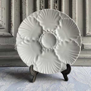 May include: A white ceramic oyster plate with six shell-shaped compartments and a central well, displayed on a black metal stand. The plate has a textured, ridged design, perfect for serving seafood. The background features a distressed white wooden door.