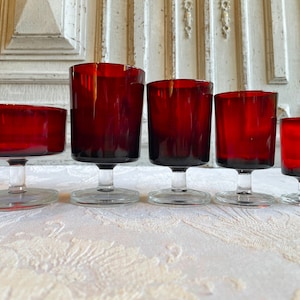 Può includere: Una collezione di cinque calici rossi rubino di diverse altezze. I bicchieri hanno una forma quadrata e poggiano su basi rotonde e trasparenti. Disposti su una tovaglia bianca fantasia.