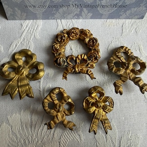 Op de afbeelding: Vijf antieke goudkleurige decoratieve elementen. De collectie bevat drie strikvormige ornamenten en een kransvormig stuk met rozen. De stukken zijn gemaakt van metaal en hebben een vintage esthetiek.