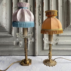 May include: Two table lamps with brass bases and clear glass bobeches featuring hanging crystals. The left lamp has a pale pink fabric shade with blue fringe trim. The right lamp has a golden-yellow fabric shade with matching yellow fringe trim.