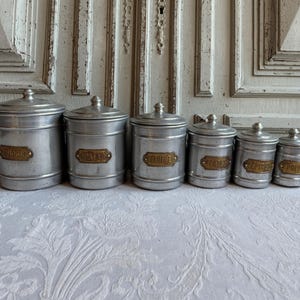 Peut inclure: Ensemble de six boîtes métalliques vintage avec couvercles, disposées en rangée. Chaque boîte argentée porte une petite étiquette dorée avec du texte. Elles reposent sur une surface blanche à motifs.