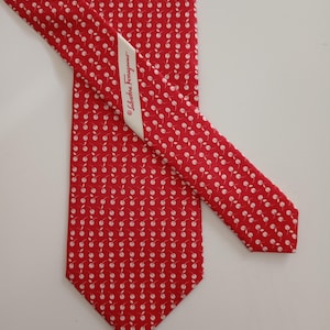 以下が含まれることがあります： 赤色のシルクネクタイ。白いサクランボの模様が繰り返されています。「Salvatore Ferragamo」と書かれたラベルが付いています。フォーマルまたはビジネスシーンに最適なクラシックなアクセサリーです。