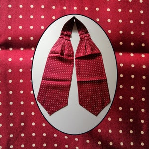 May include: A burgundy ascot with a white polka dot pattern. The ascot is made of a silky material and is folded in a classic style. The background is a matching burgundy fabric with white polka dots, creating a cohesive look.