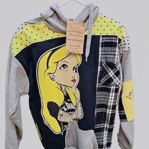 May include: A patchwork hoodie featuring a graphic of a blonde character with tattoos on a black background. The garment includes grey sleeves, yellow panels with black dots, and a black and white plaid pocket. A tag reads 1 of 1 Upcycled Hoodie.