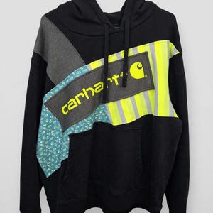 May include: A black hooded sweatshirt featuring a patchwork design across the chest. The panel includes a dark gray section with the yellow text Carhartt, a blue floral fabric segment, and a bright yellow and gray reflective striped section.