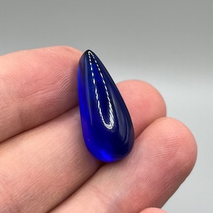 May include: A teardrop-shaped cabochon gemstone, cut and polished to a smooth, glossy finish. The gemstone is a deep, rich blue color.