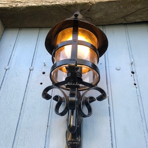 May include: An ornate, black metal outdoor wall lantern with a warm, glowing light. The lantern features a clear glass enclosure and decorative scrollwork. It is mounted on a light blue wooden surface.