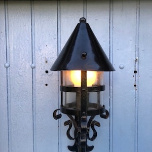 May include: Black outdoor wall-mounted lantern with a conical top and clear glass panels. The lantern has a decorative wrought iron frame with scrollwork and a warm light emanating from within. It is mounted on a light blue wooden wall.