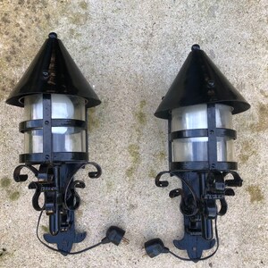 May include: Two black outdoor wall-mounted lamps with a conical top and clear glass panels. The lamps have decorative scrollwork and are connected to electrical plugs. The lamps are designed for exterior use.
