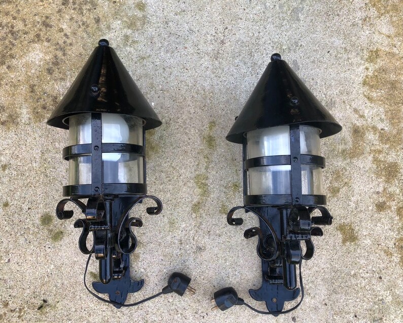 May include: Two black outdoor wall-mounted lamps with a conical top and clear glass panels. The lamps have decorative scrollwork and are connected to electrical plugs. The lamps are designed for exterior use.