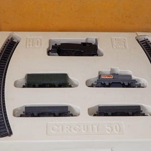 May include: A vintage HO scale model train set, featuring a black locomotive, several freight cars, and curved track pieces. The set is packaged in a white tray with the text "CIRCUIT 50". The box is yellow.