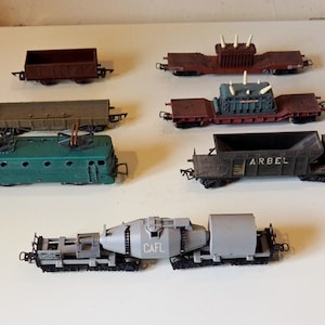 May include: A collection of model train cars in various colors and designs. Includes a green electric locomotive, several freight cars, and a specialized car with the text "CAFL". The cars are arranged on a white surface.