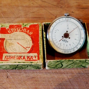 Può includere: Regolo calcolatore circolare vintage con quadrante bianco e segni neri, esposto nella sua scatola originale. La scatola è rossa con testo bianco e un diagramma. Il regolo è in una scatola verde e bianca, e il quadrante ha un'ago rosso.