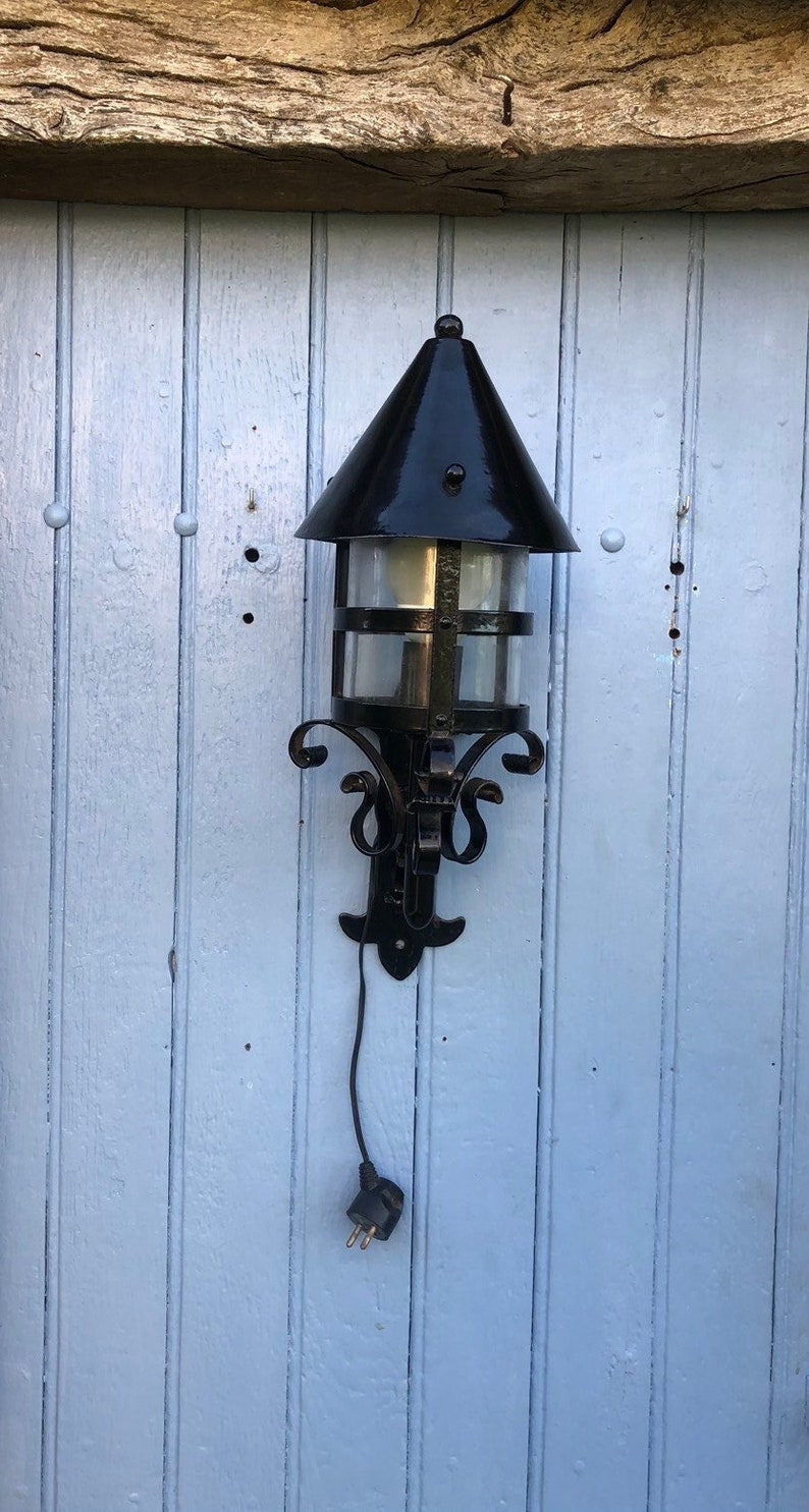 May include: Black outdoor wall-mounted lantern with a conical top and clear glass panels. The lantern has decorative scrollwork and a black electrical cord with a plug. The lantern is mounted on a light blue wooden door.