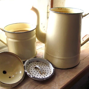 Könnte beinhalten: Eine vintage Emaille-Kaffeekanne mit cremefarbener Oberfläche und schwarzem Rand. Die Kanne hat einen Griff und einen Ausguss. Dazu gehört ein passender cremefarbener Emaille-Kaffeefilter mit schwarzem Rand und Griff. Der Filter hat einen abnehmbaren Deckel mit einem Loch in der Mitte. Die Kaffeekanne und der Filter stehen auf einer Holzoberfläche.