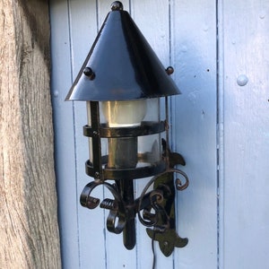 May include: Black outdoor wall-mounted lantern with a conical top and clear glass panels. The lantern has decorative scrollwork and is attached to a light blue wooden wall. A power cord extends from the bottom.