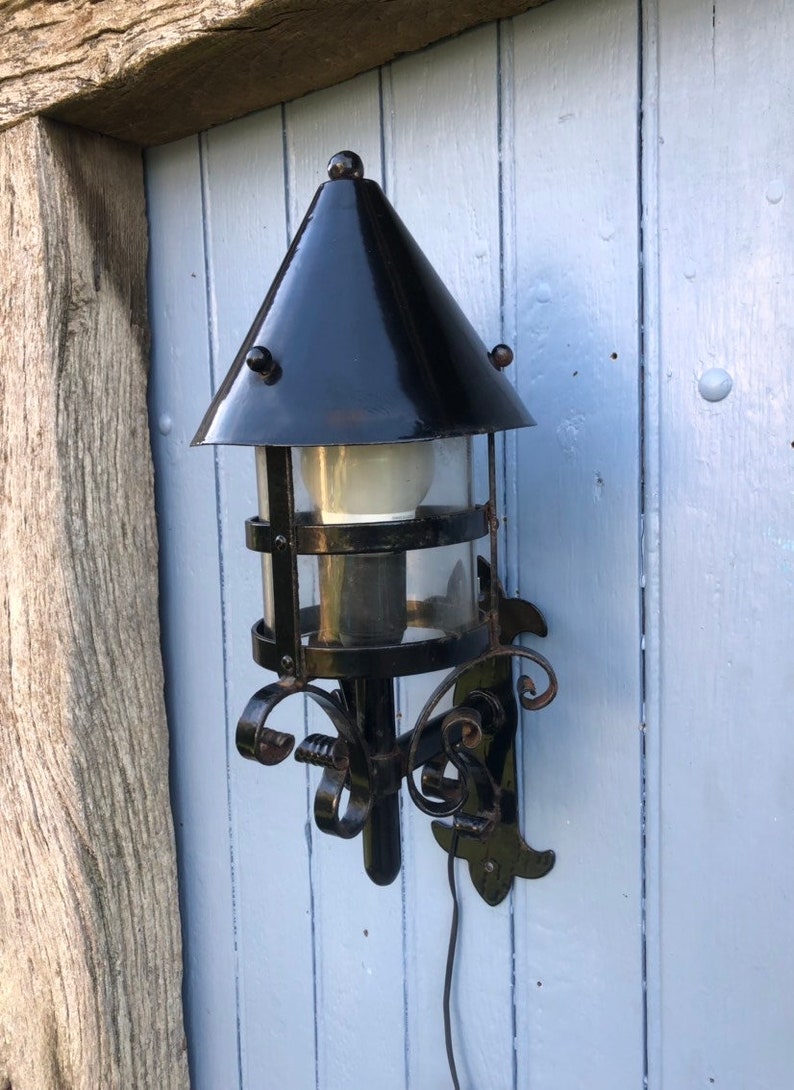 May include: Black outdoor wall-mounted lantern with a conical top and clear glass panels. The lantern has decorative scrollwork and is attached to a light blue wooden wall. A power cord extends from the bottom.