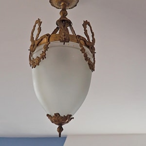 May include: An antique ceiling light fixture with a frosted glass shade and ornate gold-colored metal detailing. The fixture has a teardrop shape and is suspended from the ceiling. The metalwork includes floral and leaf motifs.