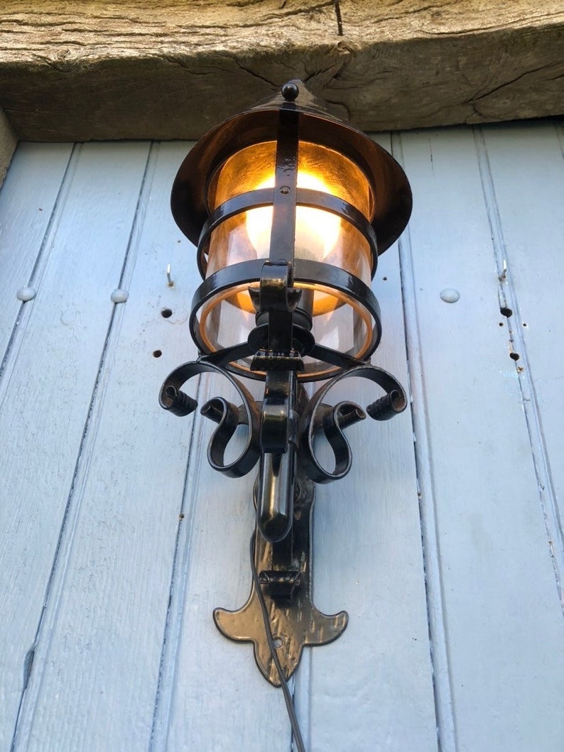 May include: An ornate, black metal outdoor wall lantern with a warm, glowing light. The lantern features a clear glass enclosure and decorative scrollwork. It is mounted on a light blue wooden surface.