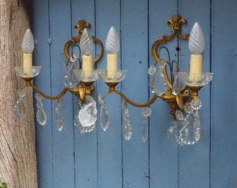 May include: Two antique gold-toned wall sconces with clear crystal accents and candle-shaped light bulbs. Each sconce has a decorative floral design and clear crystal teardrop pendants. The sconces are mounted on a blue wooden wall.