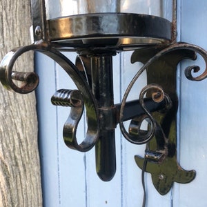 May include: Black wrought iron wall sconce with a clear glass globe. The sconce features decorative scrollwork and a bulb inside the glass. The sconce is mounted on a light blue wooden surface.