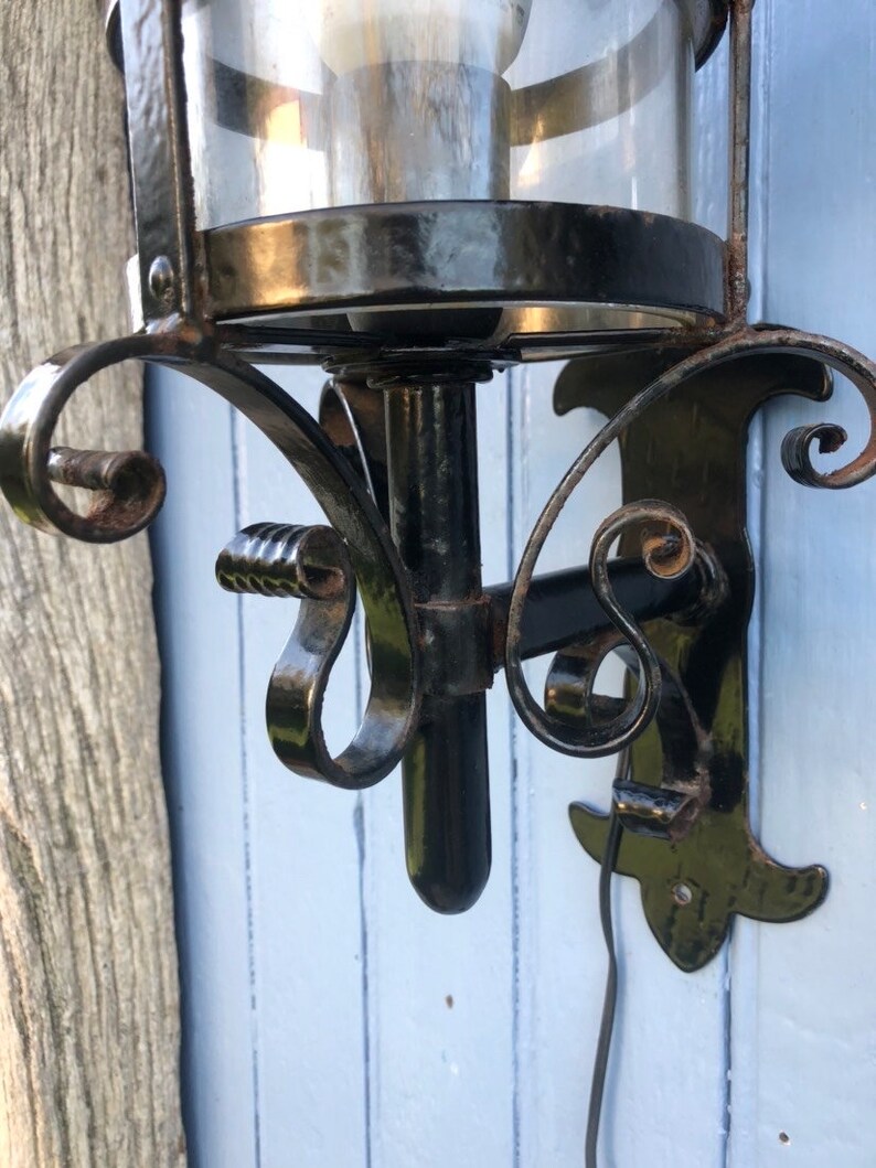 May include: Black wrought iron wall sconce with a clear glass globe. The sconce features decorative scrollwork and a bulb inside the glass. The sconce is mounted on a light blue wooden surface.