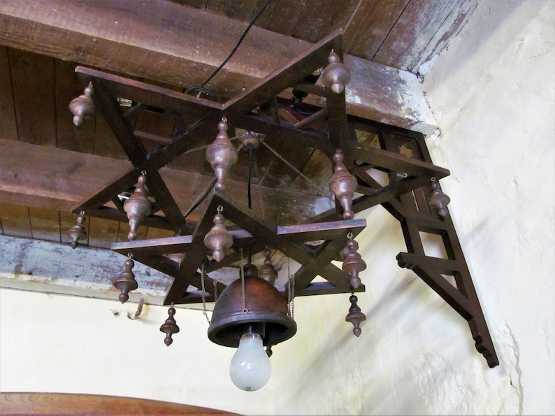 May include: A wooden star-shaped chandelier with a bell-shaped light fixture and multiple dangling wooden ornaments. The chandelier is mounted to a wooden beam.