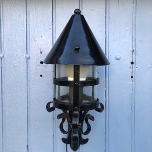 May include: Black outdoor wall-mounted lantern with a conical top and clear glass panels. The lantern has a decorative wrought iron frame with scrollwork and a bulb inside. The lantern is mounted on a light blue wooden wall.