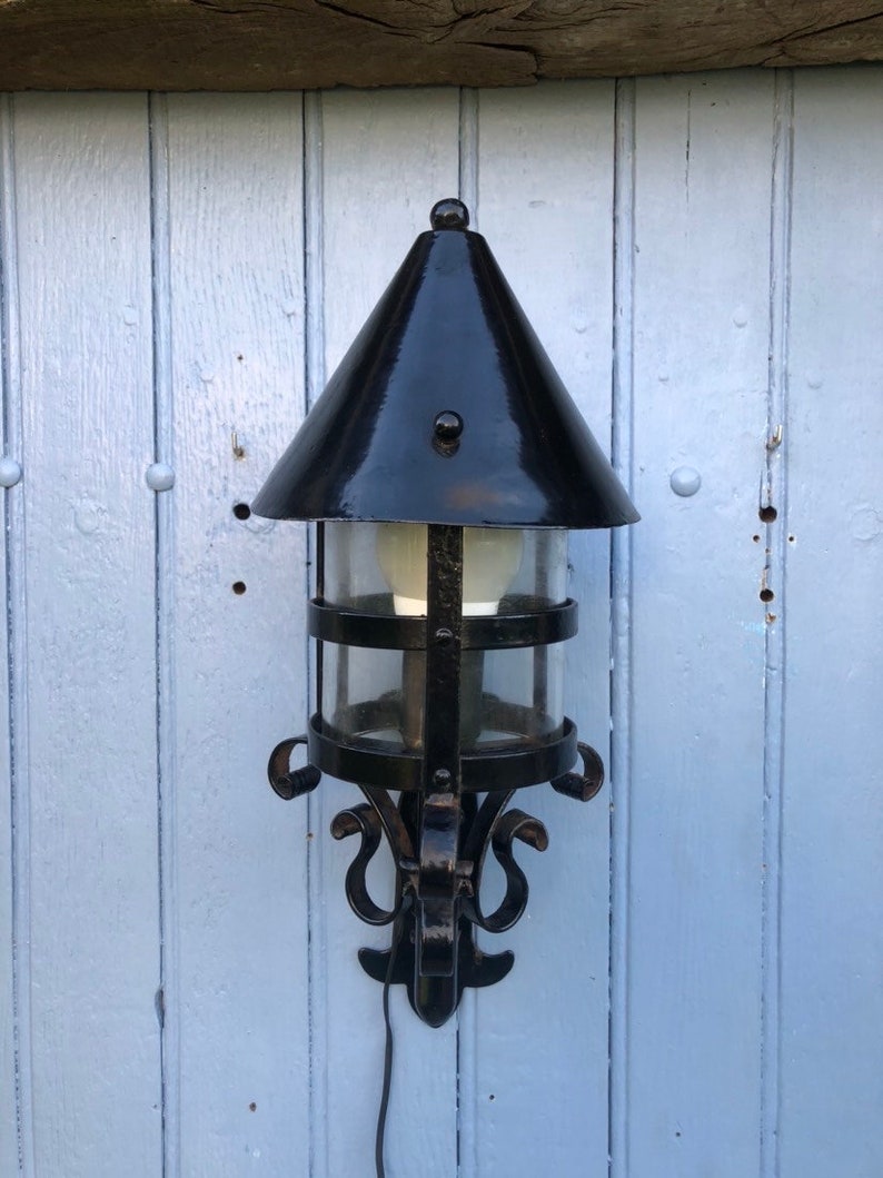 May include: Black outdoor wall-mounted lantern with a conical top and clear glass panels. The lantern has a decorative wrought iron frame with scrollwork and a bulb inside. The lantern is mounted on a light blue wooden wall.