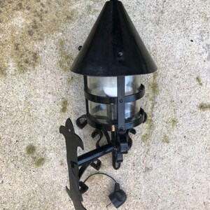 May include: Black outdoor wall-mounted lantern with a conical top and clear glass panels. The lantern has a decorative bracket and a plug. The fixture is designed for exterior use and has a classic, vintage style.