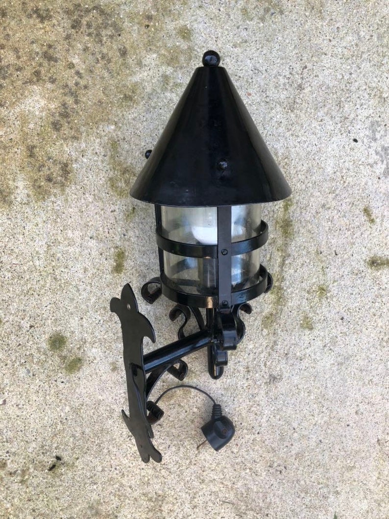 May include: Black outdoor wall-mounted lantern with a conical top and clear glass panels. The lantern has a decorative bracket and a plug. The fixture is designed for exterior use and has a classic, vintage style.
