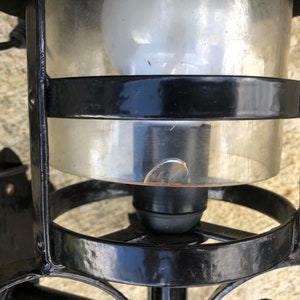 May include: Black metal outdoor lantern with a clear glass enclosure. The lantern has a bulb inside and is mounted on a wall. The glass has a small chip. The lantern is designed with decorative metalwork.