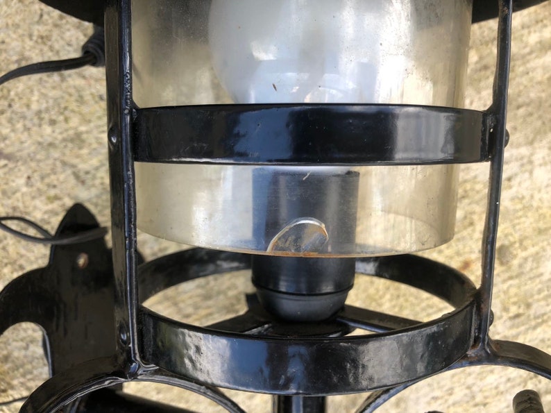 May include: Black metal outdoor lantern with a clear glass enclosure. The lantern has a bulb inside and is mounted on a wall. The glass has a small chip. The lantern is designed with decorative metalwork.