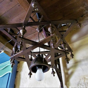 May include: A wooden star-shaped chandelier with a bell-shaped light fixture and multiple decorative finials. The chandelier is hanging from a wooden ceiling.
