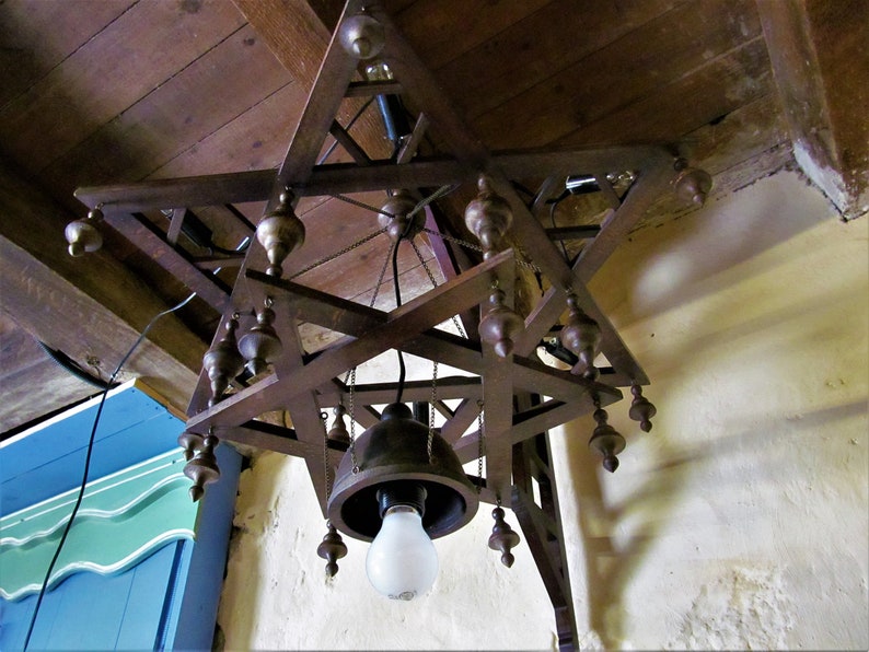 May include: A wooden star-shaped chandelier with a bell-shaped light fixture and multiple decorative finials. The chandelier is hanging from a wooden ceiling.