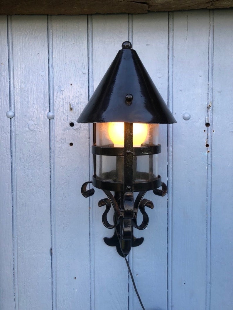 May include: Black outdoor wall-mounted lantern with a conical top and clear glass panels. The lantern has a decorative wrought iron frame with scrollwork and a warm light emanating from within. It is mounted on a light blue wooden wall.