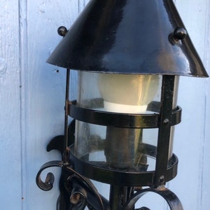 May include: Black outdoor wall-mounted lantern with a conical top and clear glass cylinder. The lantern features decorative scrollwork and a light bulb inside. The fixture is mounted on a light blue wooden surface.