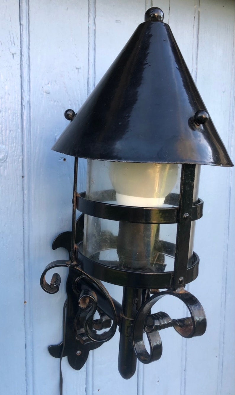 May include: Black outdoor wall-mounted lantern with a conical top and clear glass cylinder. The lantern features decorative scrollwork and a light bulb inside. The fixture is mounted on a light blue wooden surface.