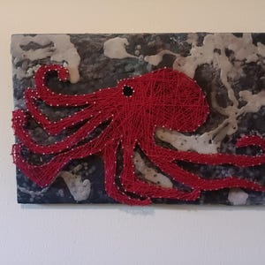 Puede incluir: Pulpo rojo de arte de hilo sobre un fondo oscuro con acentos blancos. El pulpo está hecho de hilo envuelto alrededor de clavos.