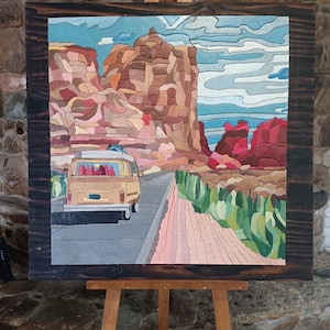 May include: A wooden framed painting of a yellow vintage van driving on a road through a desert landscape with red rock formations in the background.