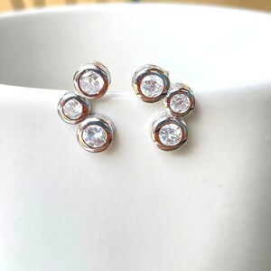 May include: A pair of silver earrings with a cluster of small, clear cubic zirconia stones. The stones are set in a circular shape and are arranged in a curved pattern.