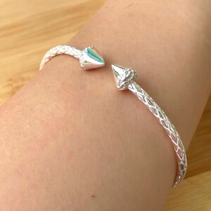 May include: A silver bracelet with a braided design and two pointed ends. The bracelet is on a person's wrist.