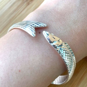 May include: Silver fish-shaped bangle bracelet with a detailed design. The bracelet is open and has a fish head on one end and a fish tail on the other.