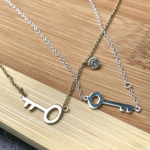 May include: Two silver key necklaces with a small diamond charm on one of the necklaces. The necklaces are on a wooden surface.