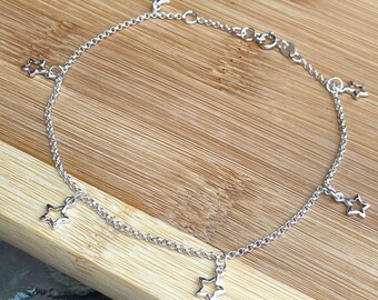 May include: A silver anklet with a delicate chain and five dangling star charms. The anklet is displayed on a wooden surface.