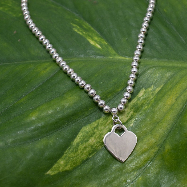 May include: A silver necklace with a heart-shaped pendant. The necklace is made of small silver beads and the pendant is a simple, flat heart.