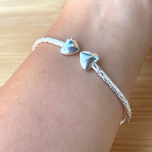 May include: A silver braided bangle bracelet with two heart-shaped charms.