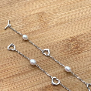 May include: Two silver bracelets with heart charms and white pearls. The bracelets are on a wooden surface.