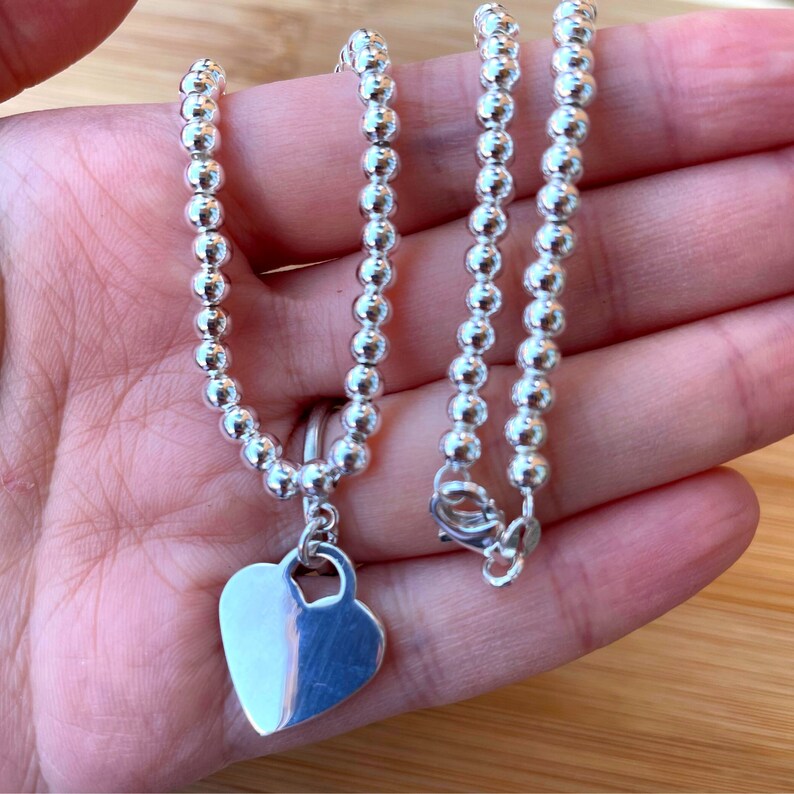 May include: A silver necklace with a heart pendant. The necklace is made of small, round beads and has a lobster claw clasp.