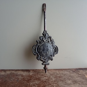 May include: An antique clock pendulum with a decorative, ornate design. The pendulum is made of dark metal and features a circular emblem with a building illustration. The pendulum has a long, slender rod with a loop at the top and a pointed tip at the bottom.